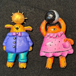 May include: Two whimsical, hand-painted figurines. One is a yellow dog with a gold sun on its head, wearing a purple coat, blue pants, and a purple bow tie. The other is an orange cat in a pink dress holding a black and white moon.