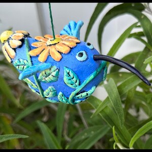 May include: A whimsical, hand-painted blue birdhouse with a long black beak, adorned with yellow and orange flowers and green leaves. The birdhouse is suspended by a green string, set against a backdrop of green foliage.