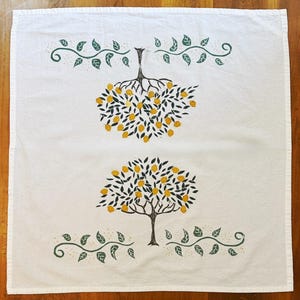 May include: White kitchen towel with a printed design of two lemon trees and leafy vines. The trees have yellow lemons and black leaves. Green and gold vines are at the top and bottom of the towel. The towel is on a wooden surface.
