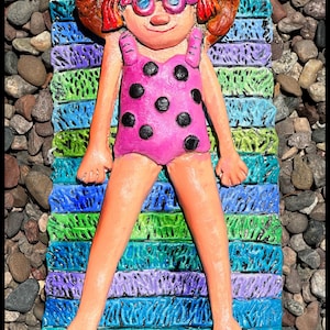 May include: A sculpted figure of a person wearing a wide-brimmed yellow hat, pink sunglasses, and a pink swimsuit with black polka dots. The figure is lying on a rectangular mat featuring horizontal stripes in shades of blue, green, and purple.
