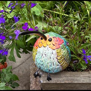 May include: A colorful, ceramic hummingbird sculpture with a long, curved beak and small black feet. The bird's body features a textured, iridescent finish with shades of green, blue, yellow, and red. It is perched on a wooden surface, surrounded by purple flowers and greenery.