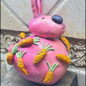 May include: A pink bunny figurine with a black nose and orange carrots attached to its body. The bunny has long ears and a smiling face. The carrots have green stems and are painted with orange and yellow details.