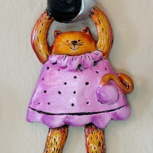 May include: A ceramic figurine of a cat wearing a pink dress with white polka dots and holding a black and silver moon above its head.