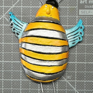 May include: A yellow and silver striped bee with blue wings and a black beret. The bee has a black nose and a small smile. The bee is standing on a gray surface.