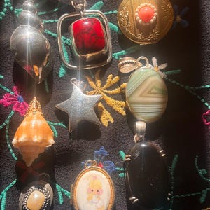 May include: A collection of vintage pendants and lockets. The pendants include a silver seashell, a red stone in a square frame, a gold locket with a coral center, a green and white stone, a silver star, a conch shell, a small locket with a painted image, and a black stone.