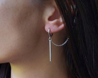May include: A silver earring with a vertical bar pendant and a delicate chain. The earring is attached to a hoop that is inserted into the ear. The earring is a simple, modern design.