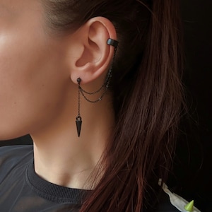 Op de afbeelding: Zwarte oormanchet en kettingoorbelset. De oormanchet wikkelt zich om de bovenkant van het oor, verbonden door delicate kettingen met een oorbel met studs en een bungelende, puntige hanger. De sieraden zijn donker, matzwart.