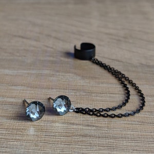 Op de afbeelding: Een set grijze kristallen oorstekers met een zwarte oormanchet verbonden door een dubbele ketting. De oorbellen hebben een rond, facetgeslepen ontwerp. De oormanchet heeft een eenvoudige, gebogen vorm. De sieraden liggen op een houten oppervlak.