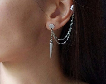 May include: Silver ear cuff and earring set. The earring features a small, round stud with a dangling chain and a cone-shaped charm. The ear cuff has a chain that connects to the earring. The jewelry is worn on the ear.