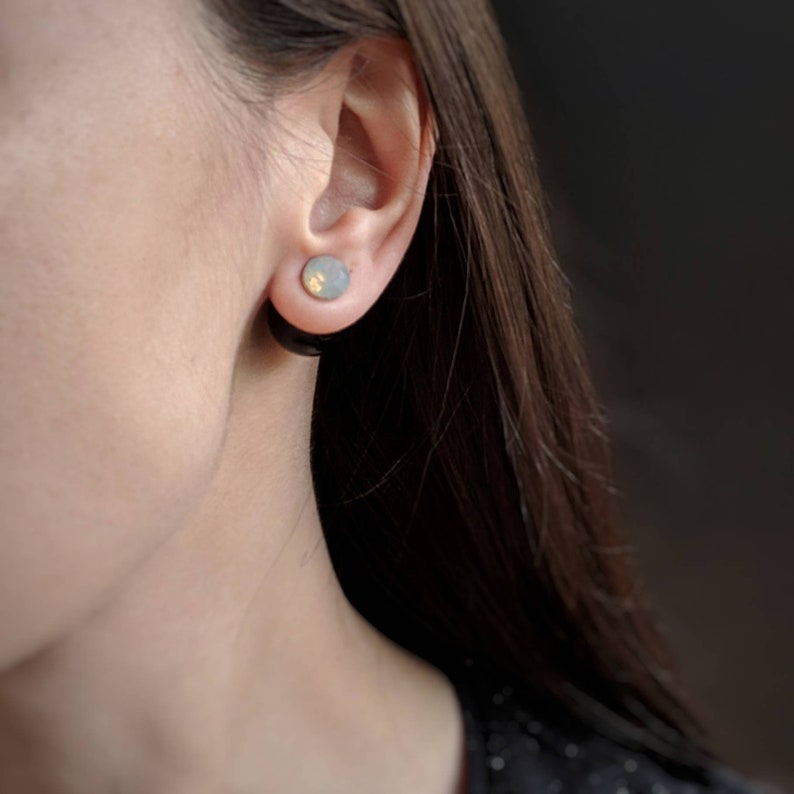 Puede incluir: Primer plano de una oreja con un pendiente redondo de color gris claro y dorado. El pendiente es de estilo tap&oacute;n, engarzado en un t&uacute;nel negro. La piel de la persona es clara y su pelo oscuro es visible.