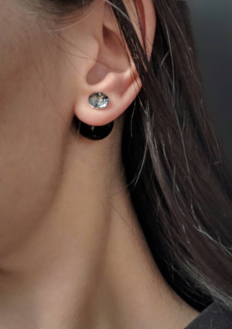 Puede incluir: Primer plano de una oreja con un par de pendientes negros y plateados. La parte delantera del pendiente es una peque&ntilde;a piedra plateada de forma ovalada. La parte trasera del pendiente es un gran perno circular negro.