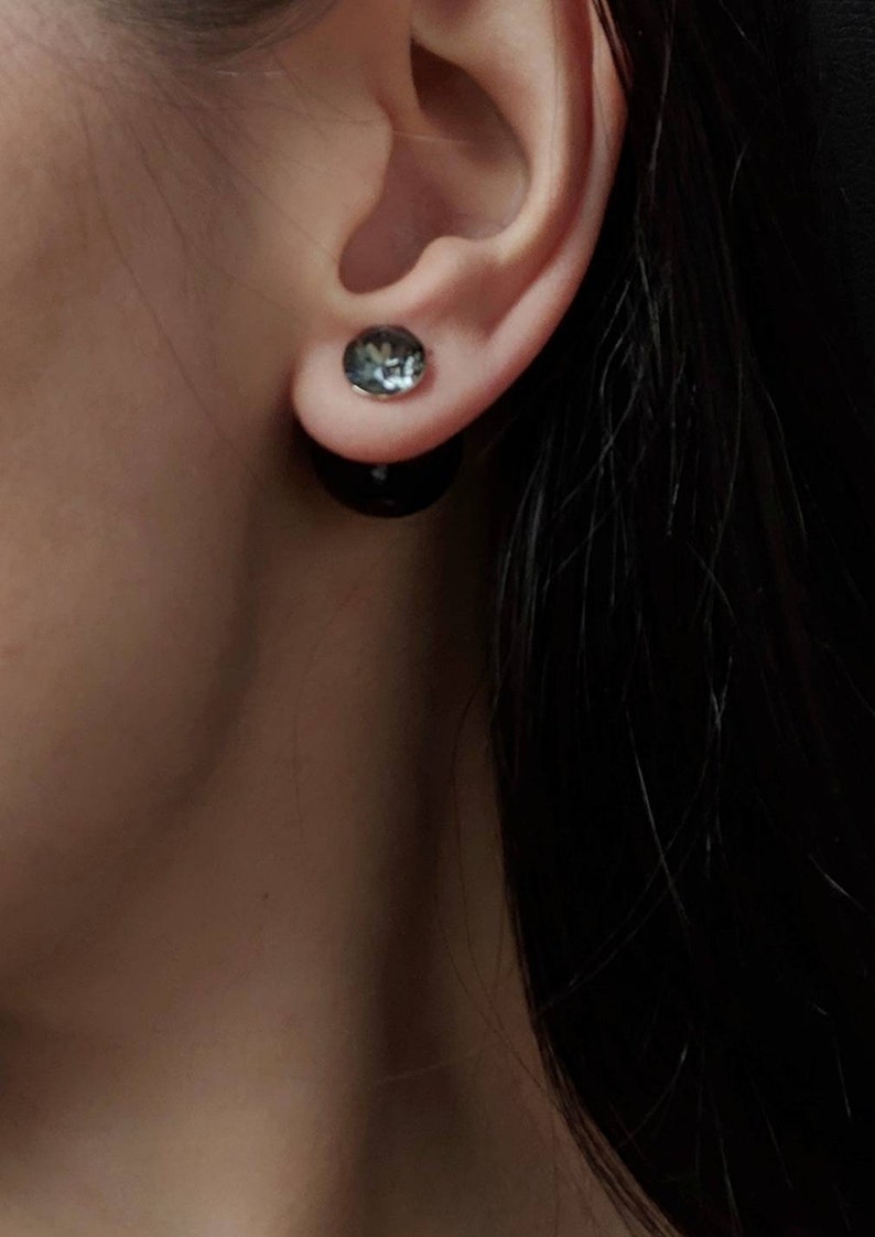 Puede incluir: Pendientes de doble cara. El perno frontal es un peque&ntilde;o cristal gris redondo, y la parte posterior es un perno negro redondo m&aacute;s grande. Los pendientes se usan en una oreja, con cabello oscuro visible.