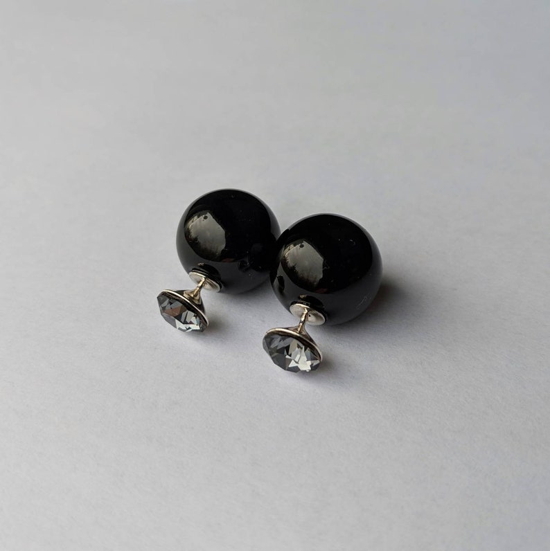 Puede incluir: Un par de pendientes esf&eacute;ricos negros y brillantes. Cada pendiente presenta una esfera negra grande y un cristal facetado plateado m&aacute;s peque&ntilde;o. Los pendientes se muestran sobre un fondo blanco liso.
