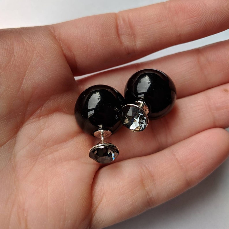 Puede incluir: Un par de pendientes negros de doble cara. Cada pendiente presenta una gran esfera negra brillante con un cristal facetado claro m&aacute;s peque&ntilde;o en el poste y la parte posterior. Los pendientes se sostienen en una mano.