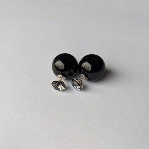 Puede incluir: Un par de pendientes negros de doble cara. Cada pendiente presenta una esfera negra grande y brillante combinada con un peque&ntilde;o y facetado cristal gris. Los pendientes tienen postes plateados y est&aacute;n sobre un fondo blanco.