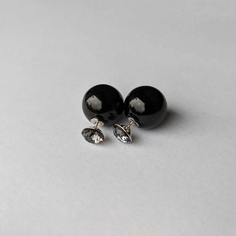 Puede incluir: Un par de pendientes negros de doble cara. Cada pendiente presenta una esfera negra grande y brillante combinada con un peque&ntilde;o y facetado cristal gris. Los pendientes tienen postes plateados y est&aacute;n sobre un fondo blanco.