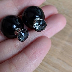 Puede incluir: Un par de pendientes negros de doble cara. Cada pendiente presenta una gran esfera negra brillante por un lado y un cristal gris facetado por el otro. Los pendientes se sostienen en una mano.