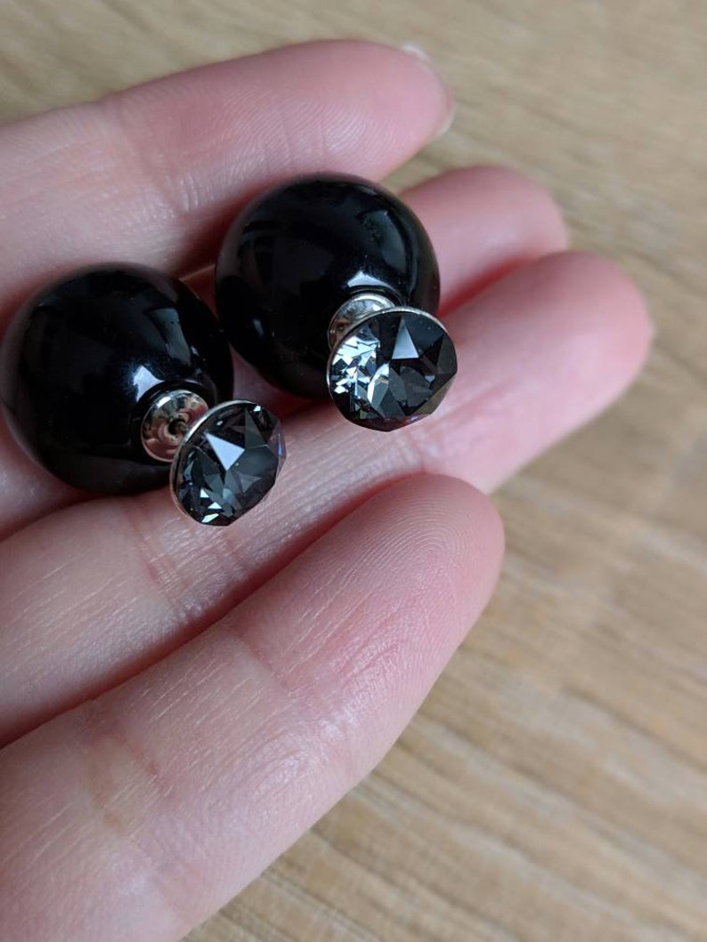 Puede incluir: Un par de pendientes negros de doble cara. Cada pendiente presenta una gran esfera negra brillante por un lado y un cristal gris facetado por el otro. Los pendientes se sostienen en una mano.