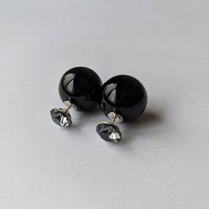 Puede incluir: Un par de pendientes negros brillantes en forma de bola, con un poste plateado y un respaldo de cristal gris facetado. Los pendientes se exhiben sobre una superficie blanca, mostrando sus cualidades reflectantes y su dise&ntilde;o elegante.