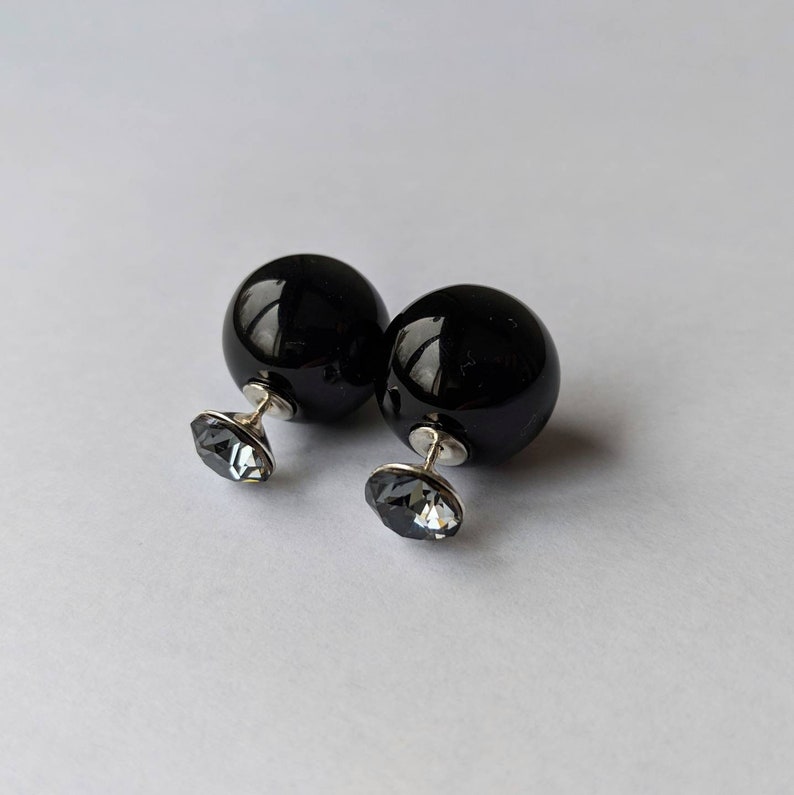 Puede incluir: Un par de pendientes negros brillantes en forma de bola, con un poste plateado y un respaldo de cristal gris facetado. Los pendientes se exhiben sobre una superficie blanca, mostrando sus cualidades reflectantes y su dise&ntilde;o elegante.
