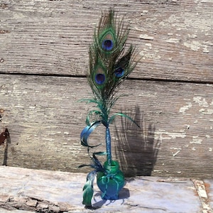 Puede incluir: Un arreglo decorativo de plumas de pavo real en una pequeña botella de vidrio de color verde azulado. Las plumas son verdes y marrones con marcas de ojos azules y negras. La botella está atada con hilo verde azulado y se encuentra sobre una superficie de madera envejecida.
