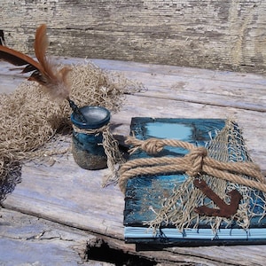 Pode incluir: Um diário azul e marrom com uma corda amarrada ao redor e um pequeno amuleto de âncora. O diário está sentado em uma superfície de madeira desgastada com uma pena de pena marrom e um frasco de vidro.