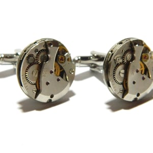 May include: A pair of silver cufflinks showcasing the detailed internal workings of a watch. The circular, metallic faces reveal gears, springs, and other components, with a polished silver backing and post.