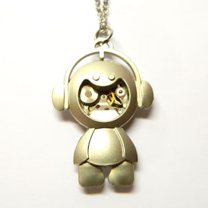 Może przedstawiać: Srebrny naszyjnik z wisiorkiem przedstawiający robota z słuchawkami. Twarz robota odsłania skomplikowane koła zębate zegarka. Wisiorek wisi na delikatnym srebrnym łańcuszku, tworząc unikalny i artystyczny dodatek.