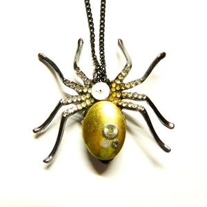 May include: A steampunk-style spider pendant necklace. The spider has a gold-colored body, rhinestone accents, and mechanical gear details. The legs are dark gray, and the chain is black.