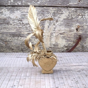 Könnte beinhalten: Eine goldfarbene, herzförmige Vase mit einem goldfarbenen Federpalmenbaum im Inneren. Die Vase und der Palmenbaum stehen auf einer verwitterten Holzoberfläche.