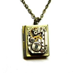 Puede incluir: Un collar dorado con un colgante de medallón que presenta un mecanismo de relojería visible. El medallón está grabado con un diseño decorativo.