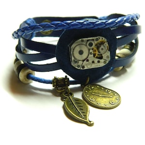 May include: A blue leather bracelet with multiple straps and a braided top layer. The bracelet features a circular navy blue suede centerpiece with a watch mechanism and two dangling charms: a leaf and a clock.
