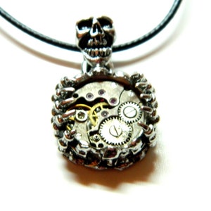 Può includere: Collana con pendente argentato a forma di teschio. Il pendente mostra gli ingranaggi interni di un orologio, con ingranaggi e gioielli visibili. La collana ha un cordino nero.