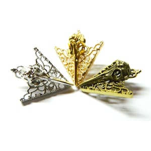 Könnte beinhalten: Drei dekorative Pfeilspitzen aus Metall mit einem Totenkopfdesign. Die Pfeilspitzen sind golden, silbern und bronzefarben.