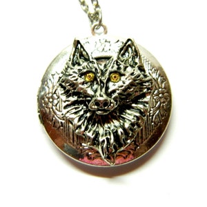 May include: Silver locket necklace with a wolf head design. The wolf has black fur and yellow eyes. The locket has a floral design around the edge.