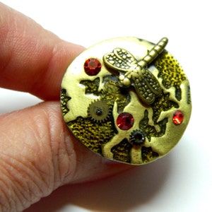 May include: A round, gold-toned metal ring with a dragonfly design. The dragonfly is made of metal and has a textured surface. The ring is decorated with red rhinestones and gears.