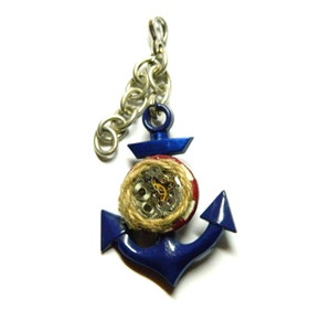 Puede incluir: Un amuleto de ancla de metal azul con un detalle de cuerda roja, blanca y marrón. El ancla tiene un diseño de relojería circular en el centro.