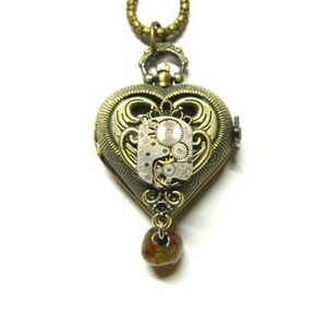 May include: A gold heart-shaped locket necklace with a steampunk design. The locket has a clear window revealing a clockwork mechanism inside. The necklace has a brown bead at the end.