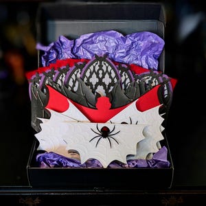 May include: A black cardboard box filled with purple tissue paper containing multiple layered paper cutouts. The designs feature black gothic arches, red bat silhouettes, and white spider web patterns with a black spider in the center.