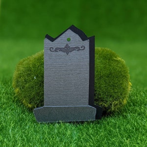 Puede incluir: Una lápida de papel gris y negro con un diseño decorativo en la parte superior. La lápida está en blanco y lista para ser personalizada.
