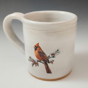 May include: A white ceramic mug with a large handle. The mug features a detailed illustration of a cardinal bird perched on a branch with red berries. The bird is primarily shades of brown and red.