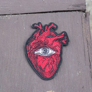 May include: A red embroidered patch featuring a stylized human heart with an eye in the center. The patch has a black border and is placed on a weathered wooden surface. The heart design has intricate details.
