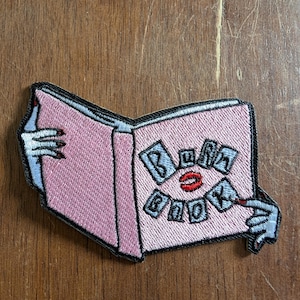 Op de afbeelding: Roze geborduurde patch in de vorm van een open boek. Het boek heeft de woorden "Burn Book" in een gestileerd lettertype. De handen die het boek vasthouden hebben rood gelakte nagels.