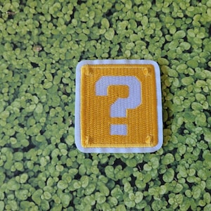 May include: A yellow question mark block patch with a white question mark. The patch is surrounded by a white border and has a stitched edge.