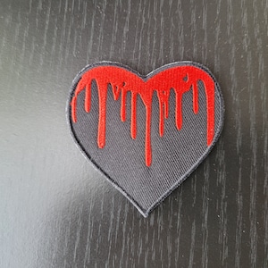 Könnte beinhalten: Schwarzer herzförmiger Patch mit roter, tropfender Farbe. Der Patch ist aus Stoff und hat einen genähten Rand.