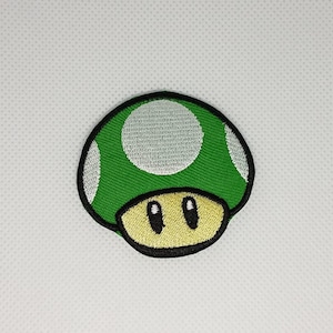 Könnte beinhalten: Gestickter Aufnäher mit einem grünen Pilz mit weißen Punkten und gelbem Sockel, schwarz umrandet. Der Aufnäher, eine beliebte Videospiel-Figur, eignet sich ideal zum Dekorieren von Kleidung oder Accessoires.