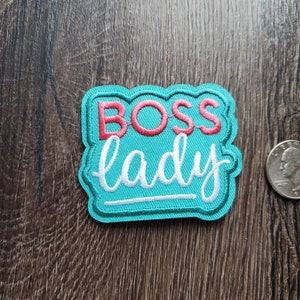 May include: A teal embroidered patch with the words "BOSS lady" in red and white script. The patch has a scalloped edge and is next to a quarter coin on a wooden surface.