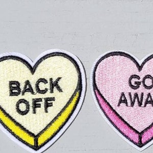 May include: Four heart-shaped iron-on patches with sarcastic messages. The patches are pink, white, light blue, and light pink. The messages are "Drop Dead", "Back Off", "Go Away", and "Beat It".