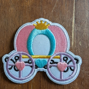 Pode incluir: Patch bordado com uma carruagem de conto de fadas rosa e azul com uma coroa dourada. A carruagem tem rodas rosa com detalhes de coração e uma carroceria azul clara. O patch é delineado em branco.