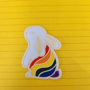 May include: White embroidered patch of a rabbit with a rainbow tail. The rainbow is made of red, orange, yellow, green, blue, indigo, and violet thread.